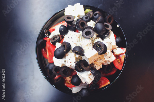 Greek salad with sirtaki cheese and black olives and a red tomato. On white cheese is ground pepper. Salad in a glass plate is on a dark table. photo