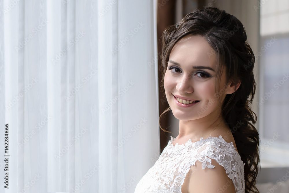 Beautiful bride stands at the window and smiles in the morning