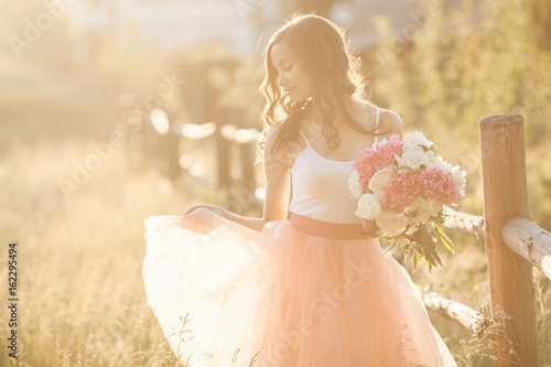 Beautiful young girl with pions on the nature otdoors in the sunshine. Pretty woman outside with flowers in the pink skirt