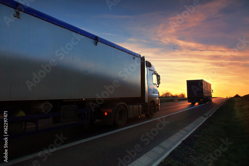 Two white truck driving through autumn landscape at sunset