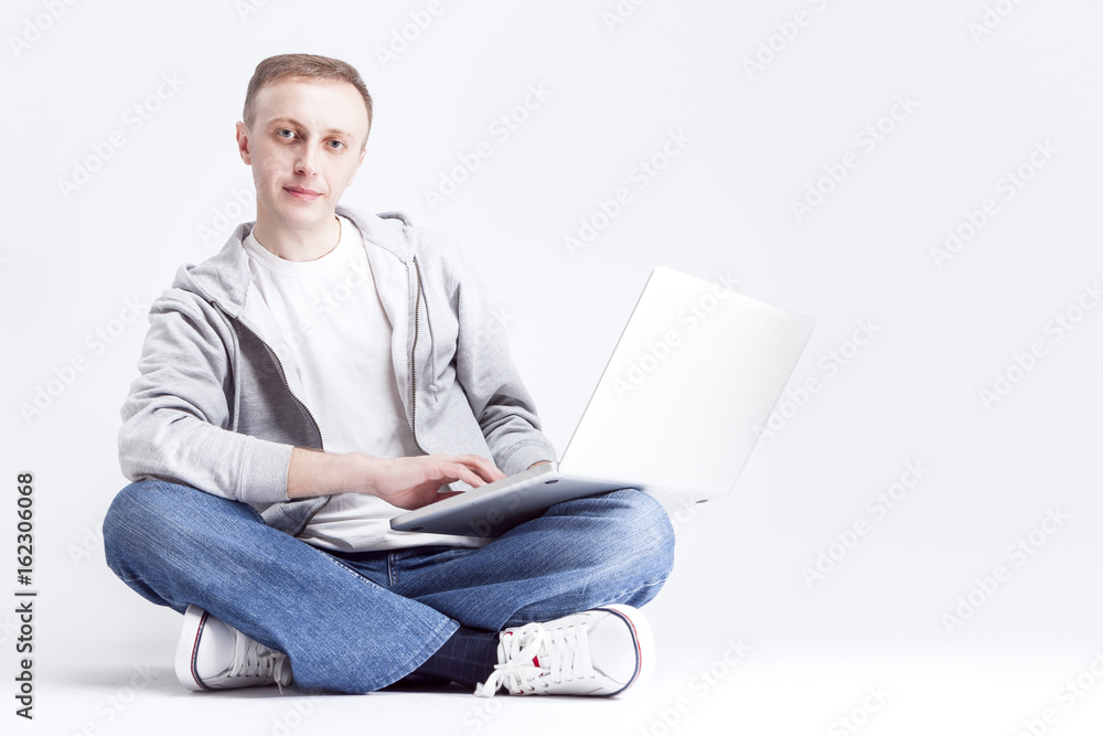 Portrait of Serious Handosme Blond Caucasian Man Sitting with Laptopp on Legs. Posing Against White Background. Portrait of Serious Handosme Blond Caucasian Man Sitting with Laptopp on Legs. Posing Against White Background.