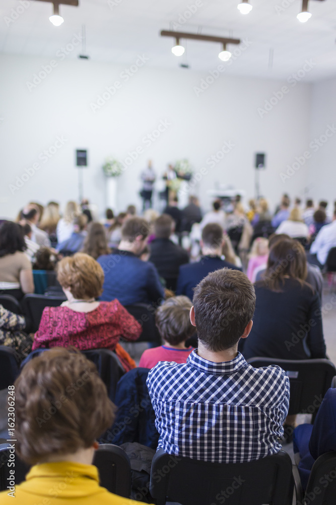 Group of People Sitting in hall During a Conference. Two Male Hosts Speaking on Stage in Front of the Audience. Group of People Sitting in hall During a Conference. Two Male Hosts Speaking on Stage in Front of the Audience.