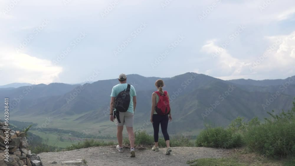 young travelers on the edge of the cliff happily put my hands up. guy and girl with backpacks in the mountains young travelers on the edge of the cliff happily put my hands up. guy and girl with backpacks in the mountains