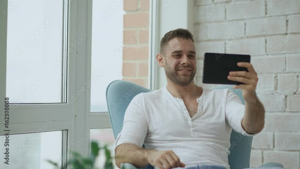 Young happy man have online video chat using digital tablet computer sitting on balcony in modern loft apartment Young happy man have online video chat using digital tablet computer sitting on balcony in modern loft apartment