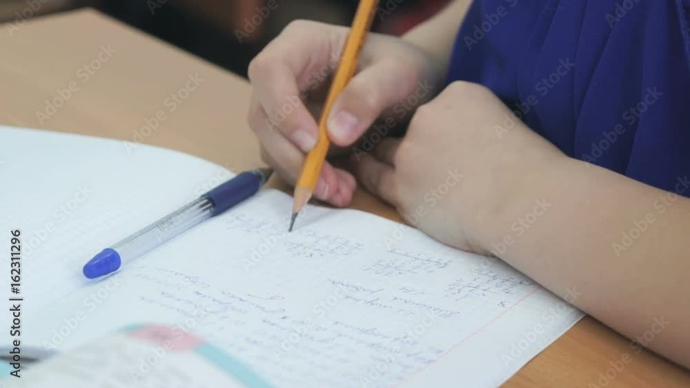 The student sits at a school desk and writes in a workbook with a ballpoint pen. Close-up The student sits at a school desk and writes in a workbook with a ballpoint pen. Close-up