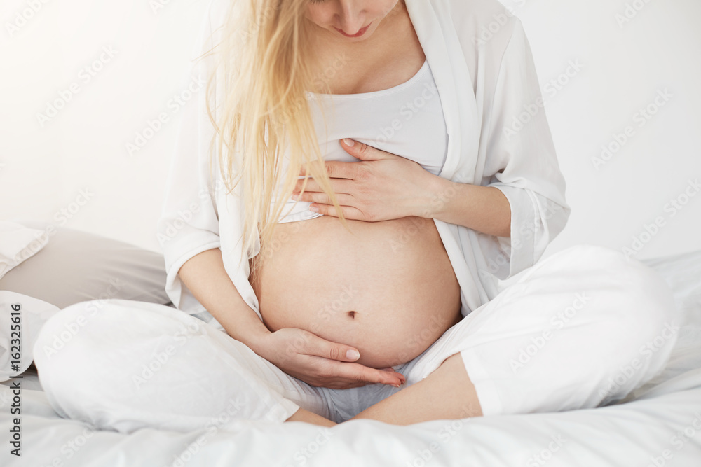 Blonde pregnant woman looking at her belly and supporting it with her hands waiting for her baby boy or girl to come. Pregnancy concept. High key. Blonde pregnant woman looking at her belly and supporting it with her hands waiting for her baby boy or girl to come. Pregnancy concept. High key.