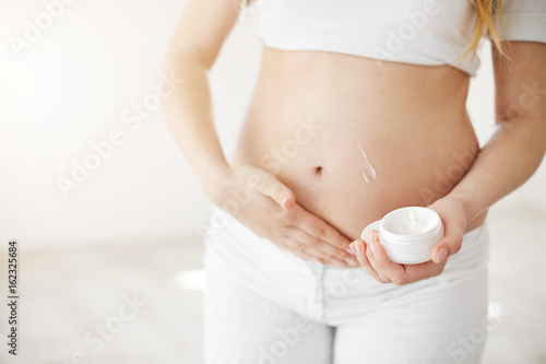 Close up of blonde early pregnant woman taking care of her belly skin using vitamin creme. Mother care concept. High key.