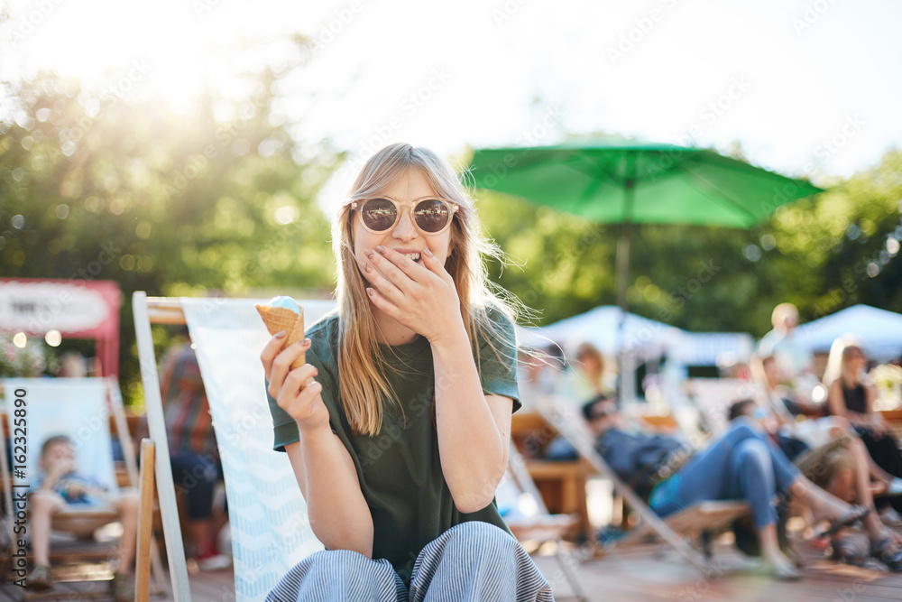 Lady eating ice cream laughing. Portrait of young female sitting in a park on a sunny day eating icecream looking at camera wearing glasses enjoying summer.