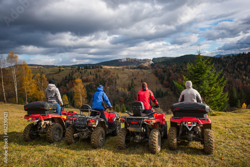Rear view of four men at quad bikes prepared to go down the hill. Beautiful landscape of rolling countryside and colorful forest under the sky with cumulus clouds in autumn