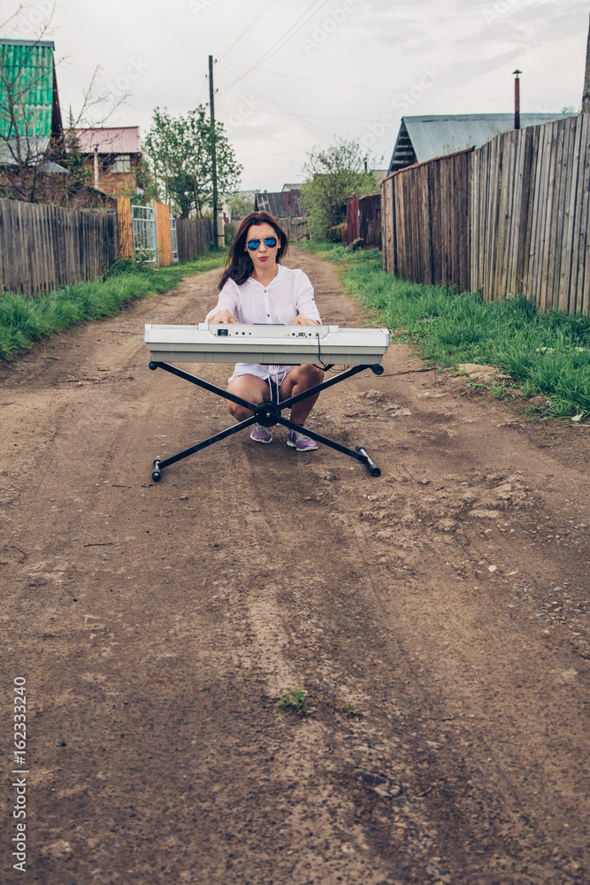 Attractive girl playing a synthesizer in nature. A woman is 25-30 years old. An electric piano stands in nature, in a field in a village. Summer day in the countryside.