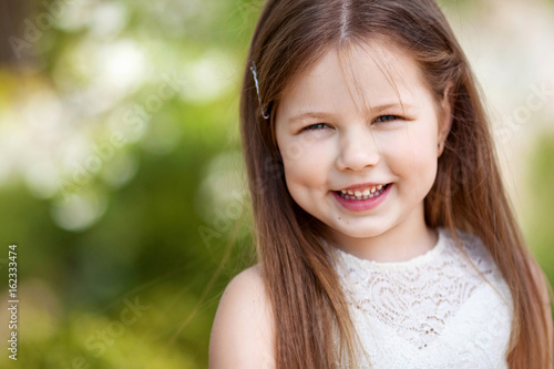 Beautiful smiling little girl in cream dress  against green of summer park