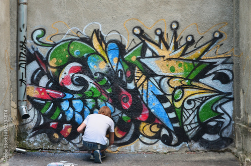 Photo in the process of drawing a graffiti pattern on an old concrete wall. Young long-haired blond guy draws an abstract drawing of different colors. Street art and vandalism concept