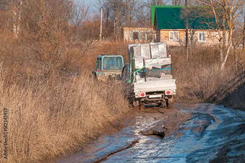 Wheeled tractor tows off-road truck with furniture settled down in mud on dirt road in village 