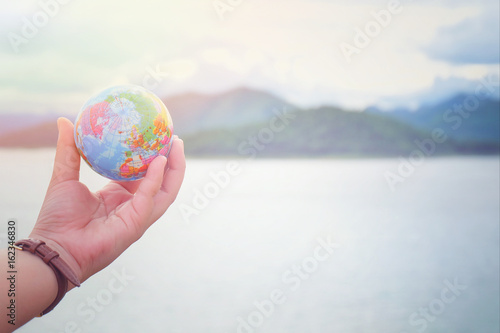 The hand holds a model globe and has a mountain and river background. Travel Concept. Communication and Technology Concept. Business Concept.