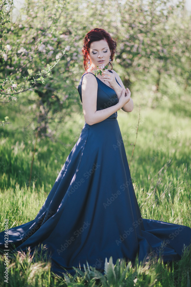 Young beautiful woman with red hair in a blue dress posing in a blooming garden Young beautiful woman with red hair in a blue dress posing in a blooming garden
