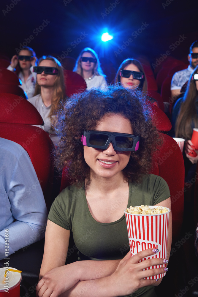 Vertical portrait of a beautiful young woman smiling enjoying 3D movie premiere at the cinema holding popcorn sitting relaxed entertaining activity spectator viewer positivity technology. Vertical portrait of a beautiful young woman smiling enjoying 3D movie premiere at the cinema holding popcorn sitting relaxed entertaining activity spectator viewer positivity technology.