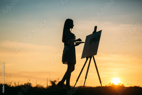 Silhouette of a girl. The blonde girl paints a painting on the canvas with the help of paints. A wooden easel keeps the picture.