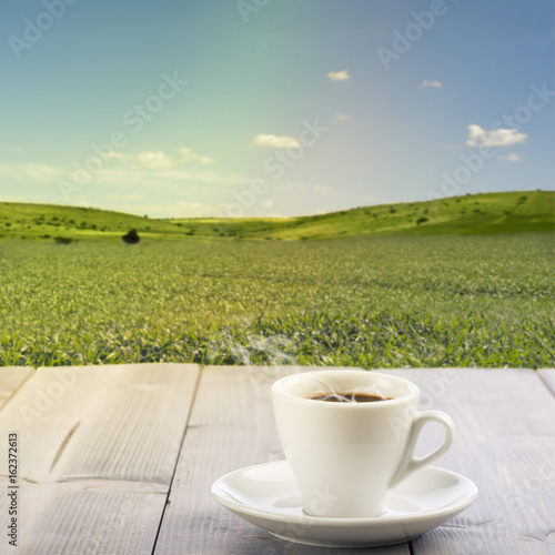 steamed cup of coffee on wooden table with sunny landscape on background.