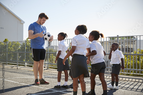 Billede på lærred Young kids in a school playground with teacher holding ball