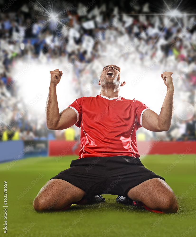 Obraz premium happy and excited football player in red jersey celebrating scoring goal kneeling on grass pitch