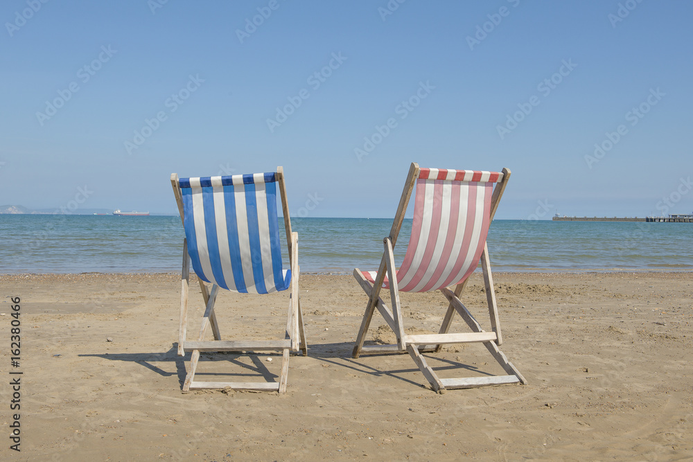 Two empty linen beach chairs one blue and one red in the middle of the image on the beach of Weymouth of the United Kingdom, facing the sea on a sunny day Two empty linen beach chairs one blue and one red in the middle of the image on the beach of Weymouth of the United Kingdom, facing the sea on a sunny day