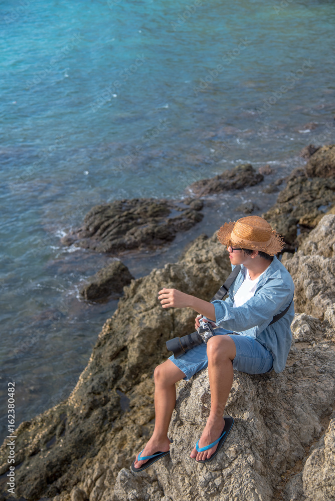 young asian man traveler sitting on the rock cliff near tropical sea, summer holiday and vacation travel concepts