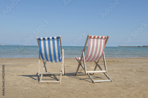 Two empty linen beach chairs one blue and one red in the middle of the image on the beach of Weymouth of the United Kingdom, facing the sea on a sunny day © Elles Rijsdijk