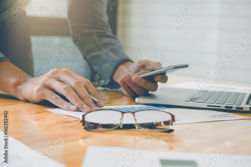 business man holding smartphone and writing financial paper work and laptop on wooden desk indoor office with morning light. Vintage filter effect.