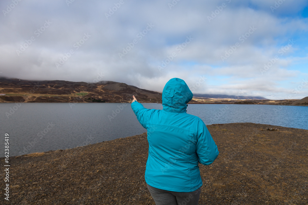 Woman in a blue jacket with her back to the photographer on the coast of huge cold lake on the stony rocky desert landscape of Iceland