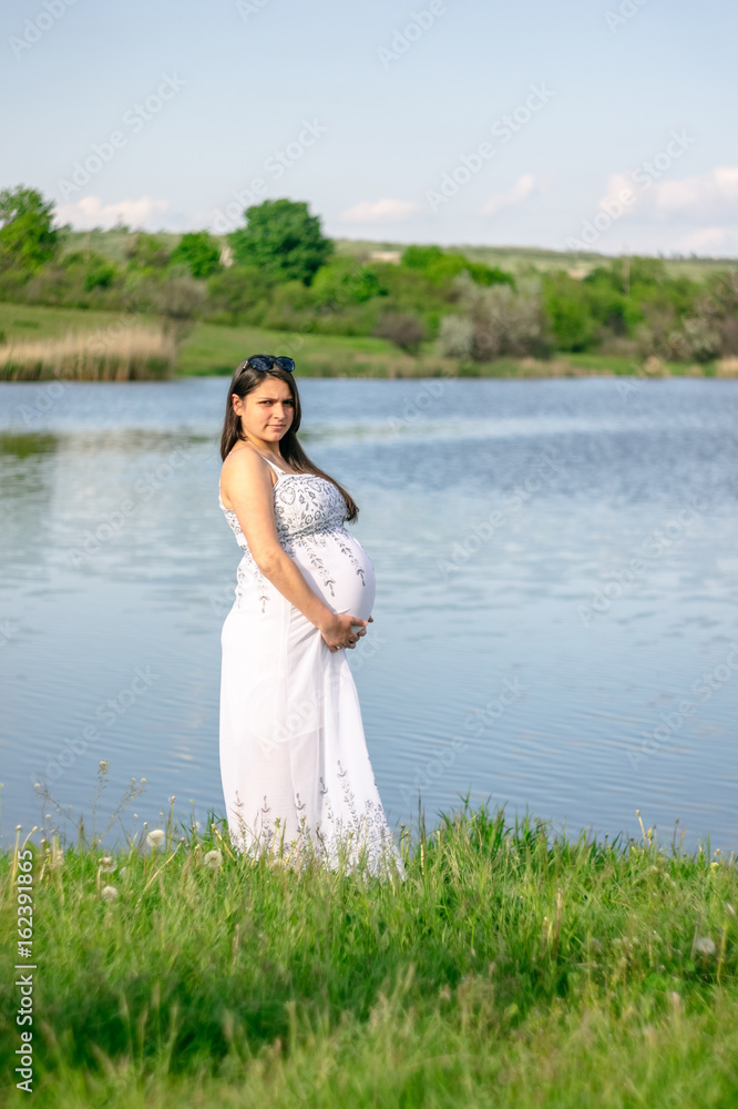 A pregnant woman is standing on the lake in a white dress. A pregnant woman is standing on the lake in a white dress.