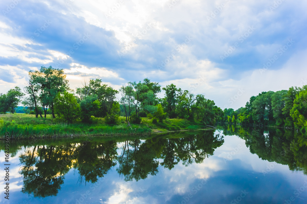 Calm beautiful scenic landscape with blue river, green trees and reflecting in water with cloudy sky. Magical sunset over the river in rural terrain. Natural, wild landscape. Calm beautiful scenic landscape with blue river, green trees and reflecting in water with cloudy sky. Magical sunset over the river in rural terrain. Natural, wild landscape.