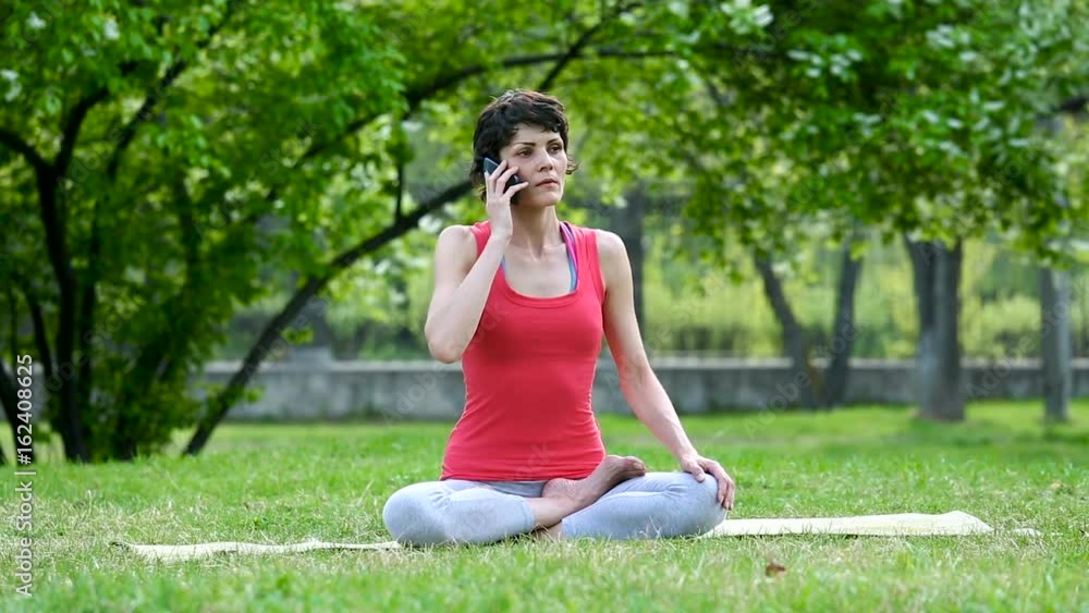 Girl is talking on a mobile phone during yoga exercise training. Happy young woman calling on smartphone and laughing sitting at yoga mat in city park. Girl is talking on a mobile phone during yoga exercise training. Happy young woman calling on smartphone and laughing sitting at yoga mat in city park.