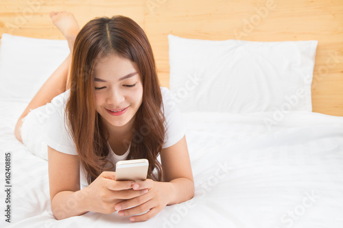happy young beautiful asian woman laying in her white and brightly lit bedroom using her smart phone
