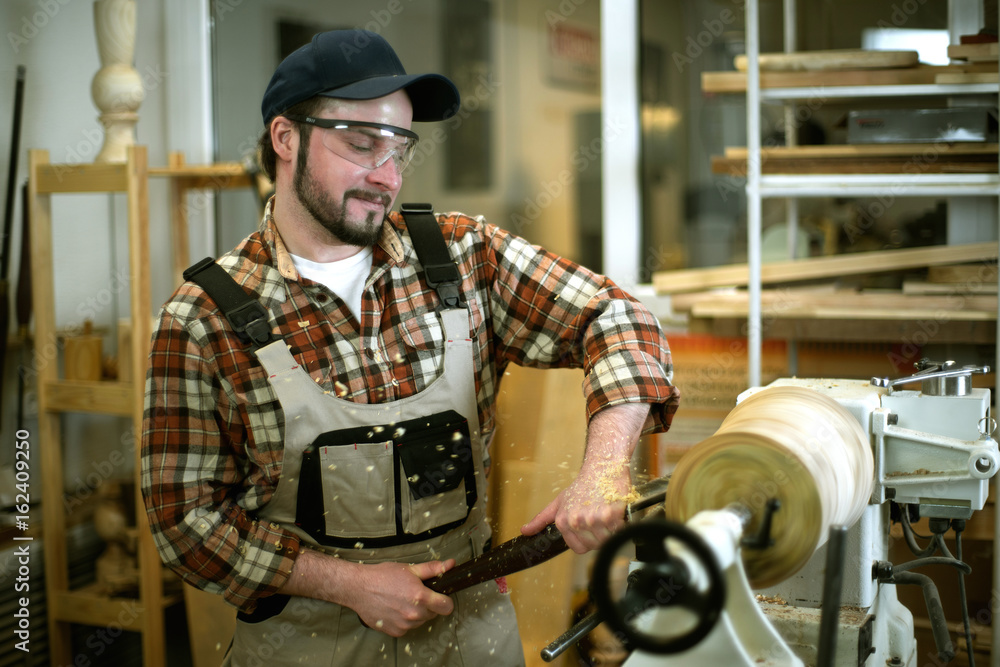 Young attractive white male in coverall and plaid shirt, working on woodworking lathe machine in the carpenter workshop Young attractive white male in coverall and plaid shirt, working on woodworking lathe machine in the carpenter workshop