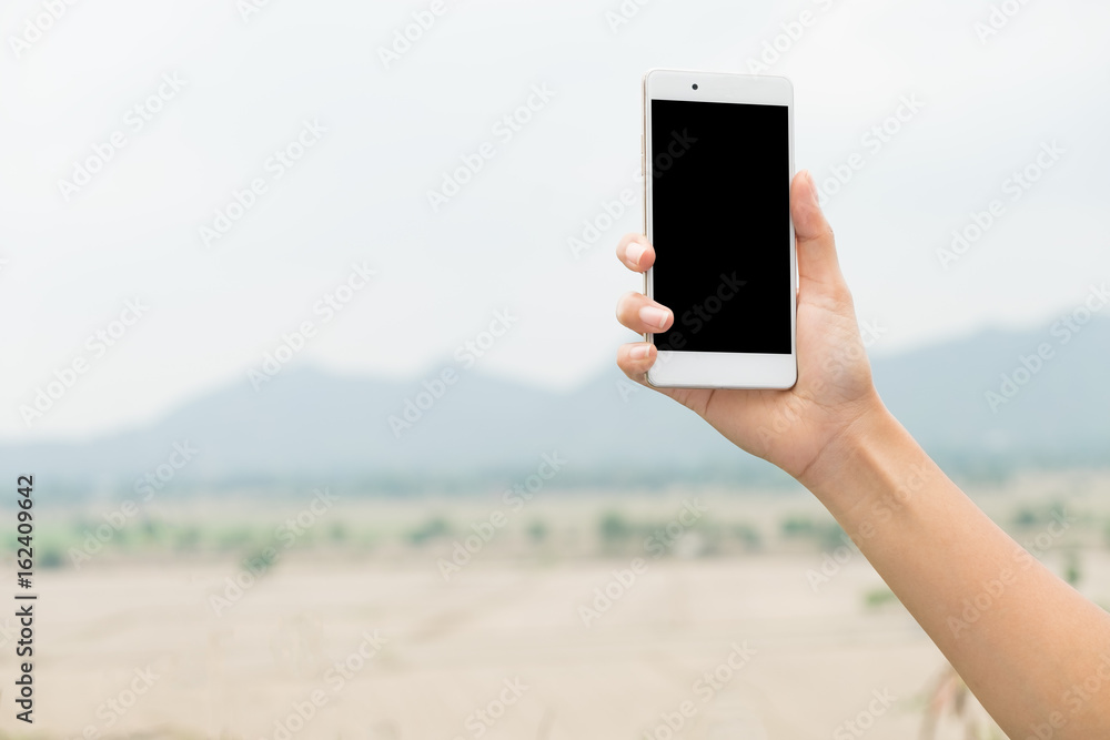 Female right hand holding and touching on mobile smartphone show back side. He is searching something. on blur forest mountain and sky background
