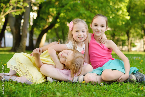 Three cute little sisters having fun together on the grass on a sunny summer day. Funny kids hanging together outdoors.