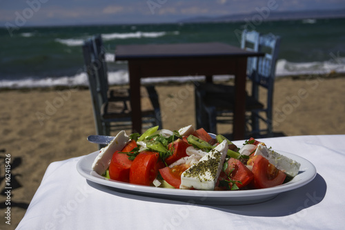 Greek salad with feta cheese  onions  olive oil and tomatoes by the sea.  Xoriatiki salad served on a table by Greek beach with blurred beach tavern table and wooden blue chairs background.