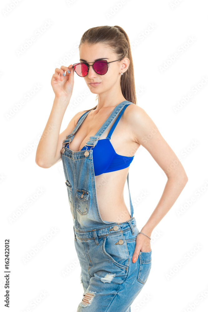 sexy young woman stands in the Studio in jeans overalls and adjusts her glasses