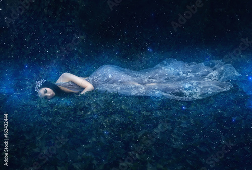 fantasy woman Sleeping Beauty. girl lies on the grass in dark, dense forest magical woods. An unusual transparent blue creatuve long dress. Artistic processing art photography. Sweet dream