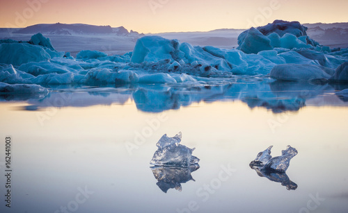 Two small pieces of ice flowing and reflecting in cold lake with a big icebergs behind, jokulsarlon glacier lagoon, Iceland