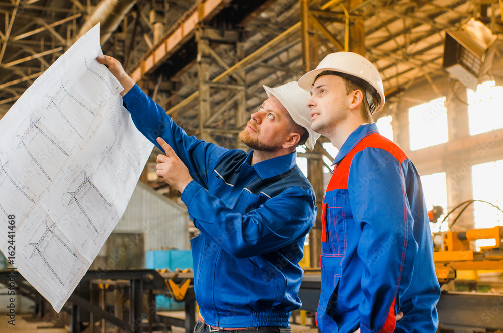 Workers in overalls and white protective helmets view the drawing on the background of an industrial metal plant. Builders in shape. Men are engineers Workers in overalls and white protective helmets view the drawing on the background of an industrial metal plant. Builders in shape. Men are engineers