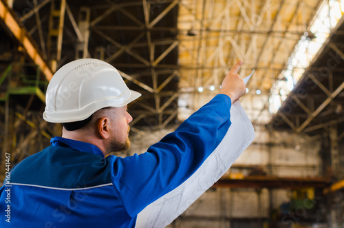 A worker in overalls and a white helmet examines the drawing and points with his index finger on the background of an industrial metallurgical plant. The builder is in shape. The man is an engineer