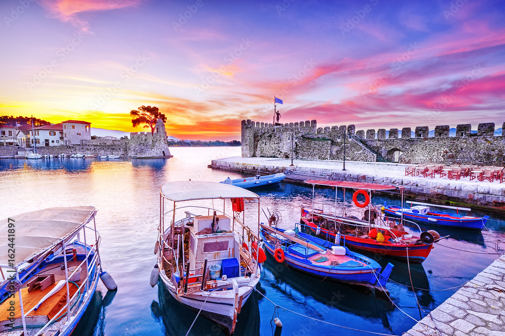Old Greek port Nafpaktos with ancient harbor walls an background and fishing boats at foreground. Epic lilac colored sunrise sky over sea scenery. Nafpaktos is famous  travel destination in Greece.
