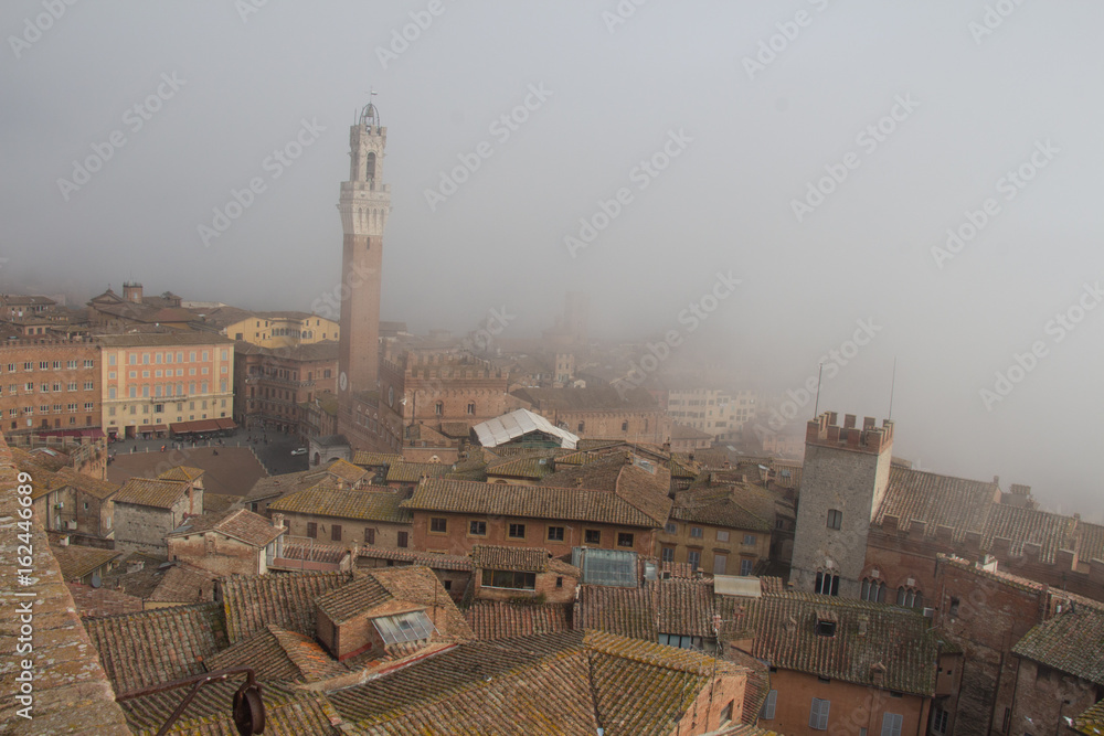 Torre del Mangia in Piazza del Campo and tupical ref roofs of Siena in mist. Tuscany, Italy.