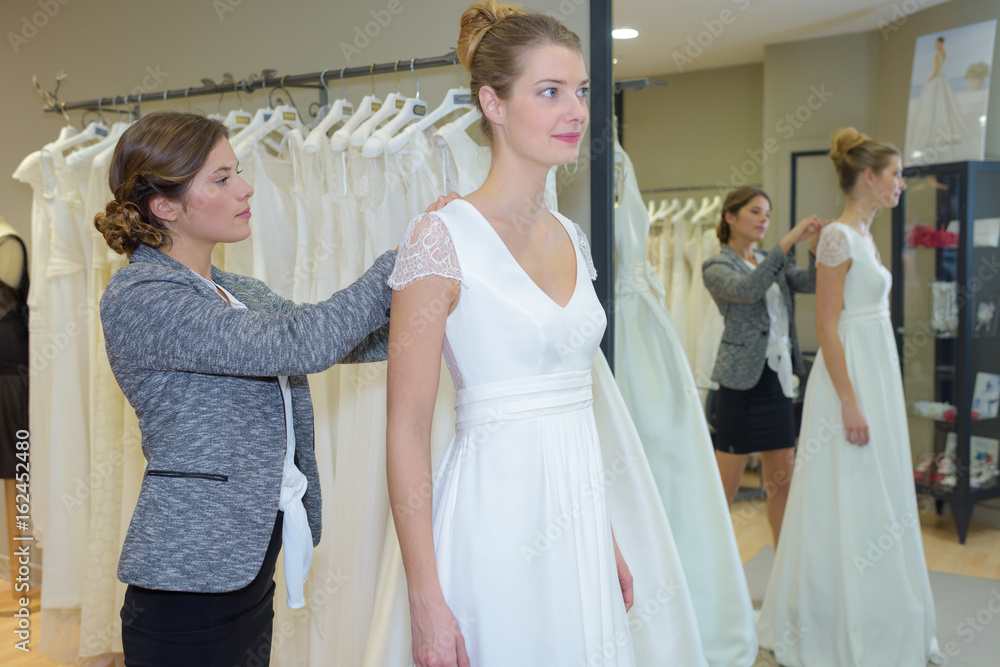 female trying wedding dress in a shop with shop assistant female trying wedding dress in a shop with shop assistant