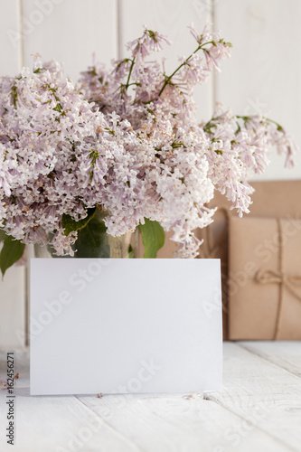 Blank white greeting card with lilac flowers bouquet and envelope on white wooden background. mock up photo