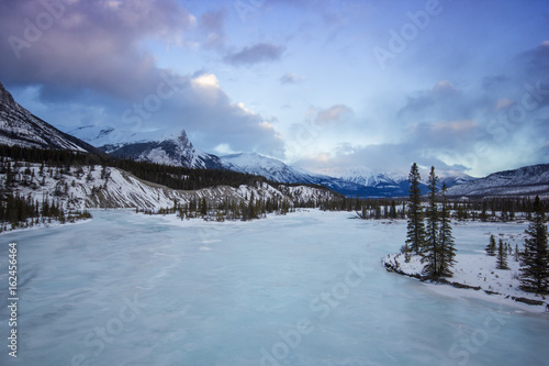 Big frozen river in mountain valley with trees and dramatic cloudy sky, Banff national park, Canada