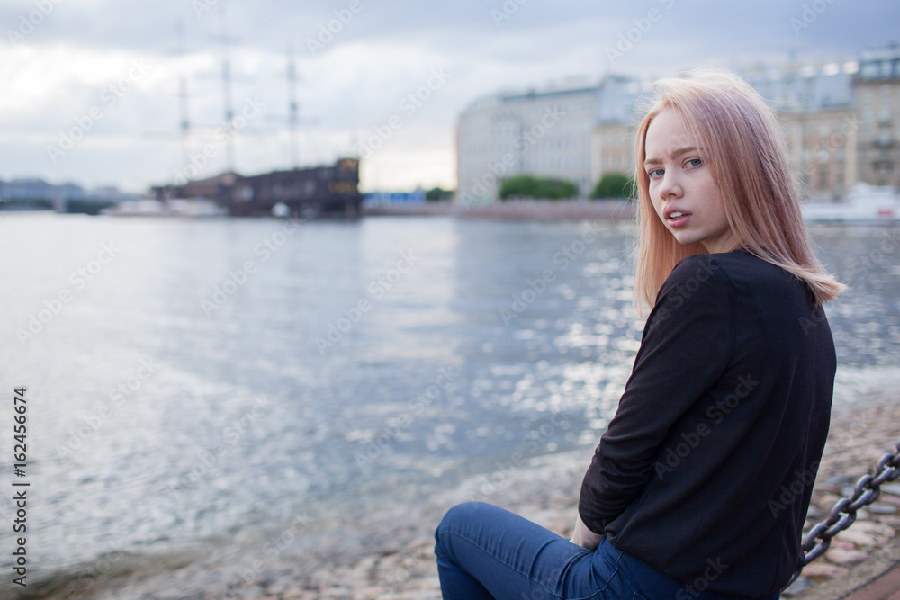 Young beautiful girl sits on the waterfront and looking at the camera. In the background is blurred old ship