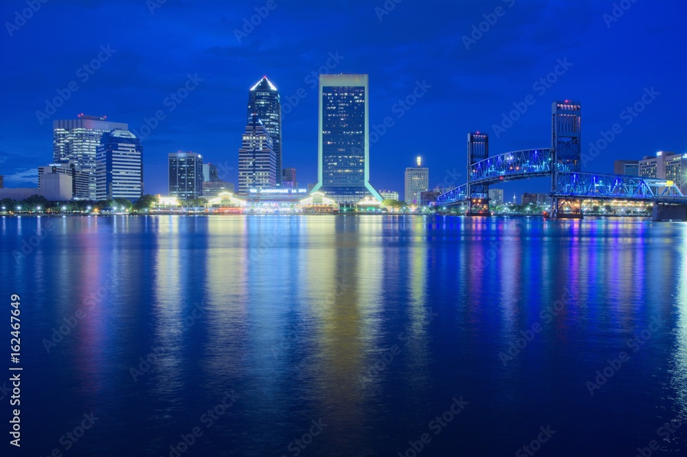 Fototapeta premium View of a downtown at dusk with buildings, bridge and colored lights reflecting over water shot as long exposure from across the water with blue sky, landscape composition