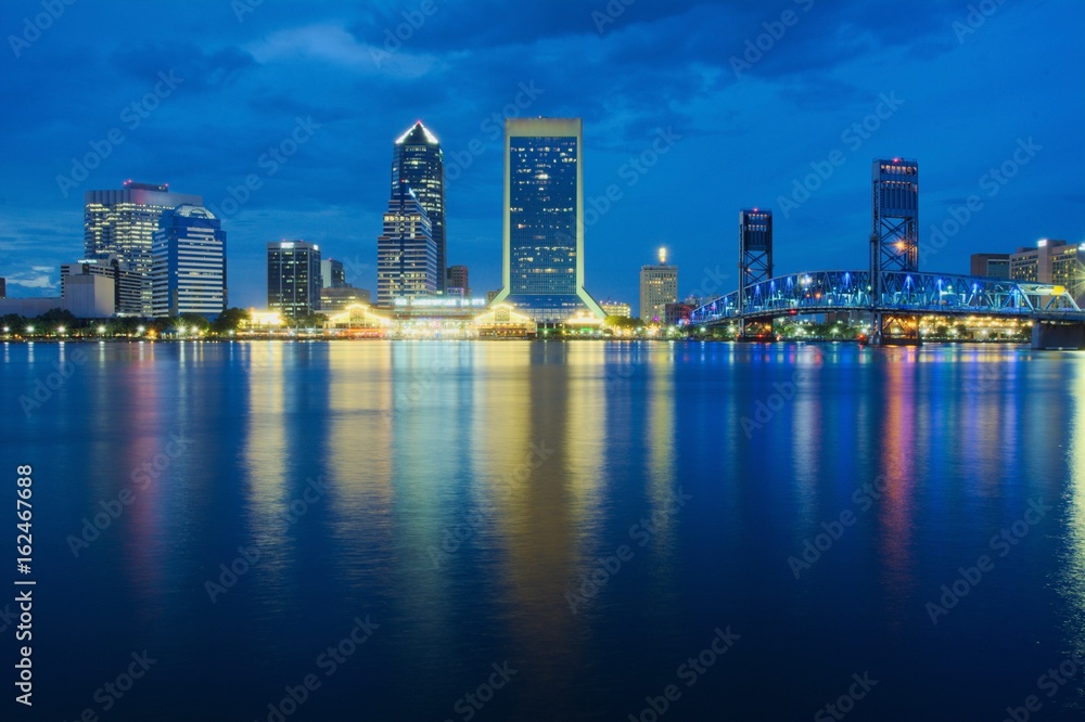 Fototapeta premium View of a downtown at dusk with buildings, bridge and colored lights reflecting over water shot as long exposure from across the water with blue sky, landscape composition